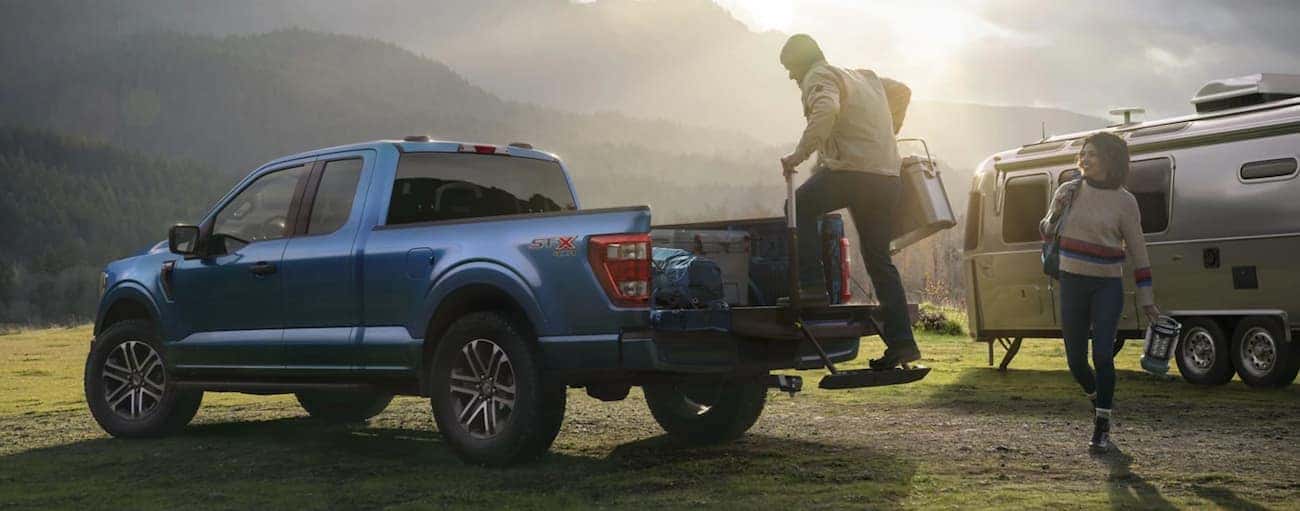 A person is loading the bed of a blue 2021 Ford F-150 using the tailgate step while parked next to an Airstream in front of mountains.
