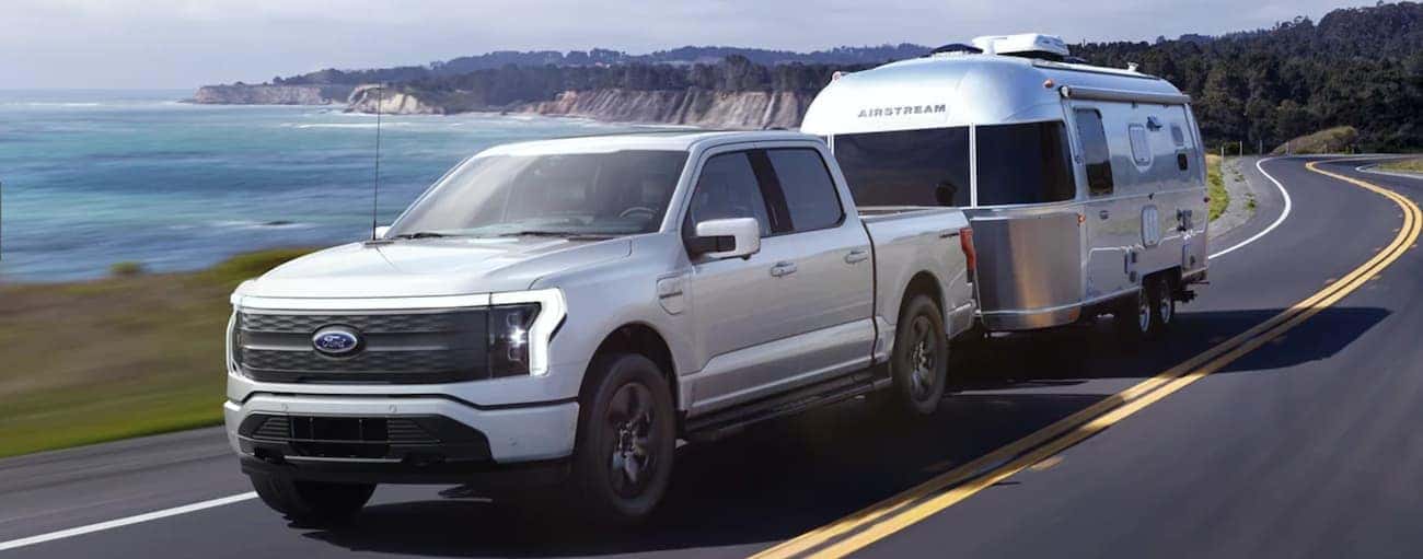 A silver 2022 Ford F-150 Lightning is towing a trailer on a road next to the ocean.
