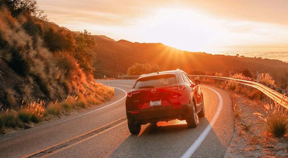 A red 2020 Mazda CX-30 is shown from the back driving along a mountain road at sunset.