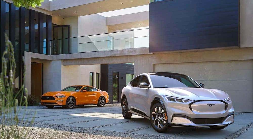 An orange 2021 Ford Mustang and a white 2021 Ford Mustang Mach-E are shown parked in a modern driveway.