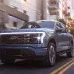 A blue 2022 Ford F-150 Lightning is shown driving through a city after leaving a Ford Lightning dealer in NY.