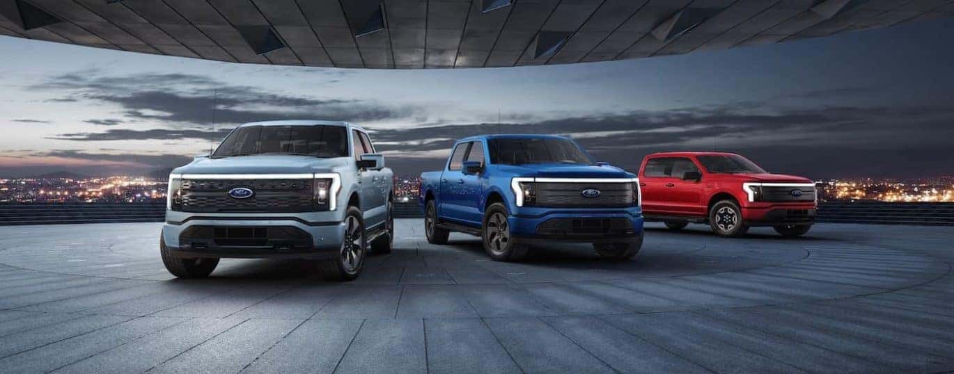 A light blue, a dark blue, and a red 2022 Ford F-150 Lightning are shown with a distant city.