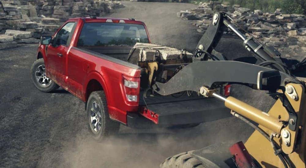 A red 2021 Ford F-150 is shown on a construction site as heavyweight machinery is loaded into the bed.