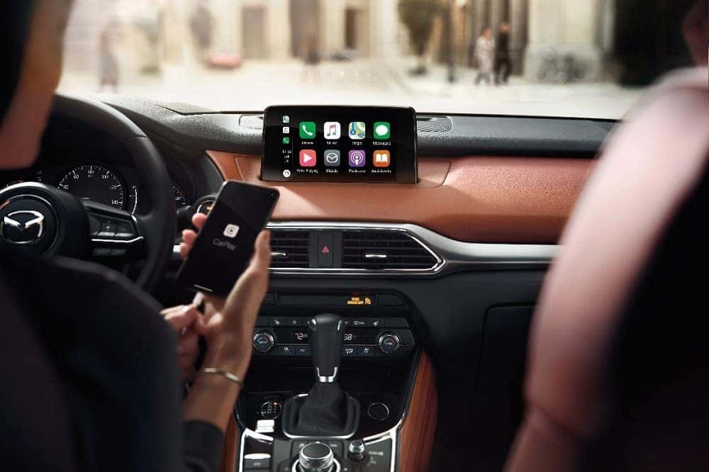 Two-tone brown and black interior of a 2019 Mazda CX-9 showcasing the MAZDA CONNECT infotainment center with Apple CarPlay for iPhone and a woman holding a black iPhone®