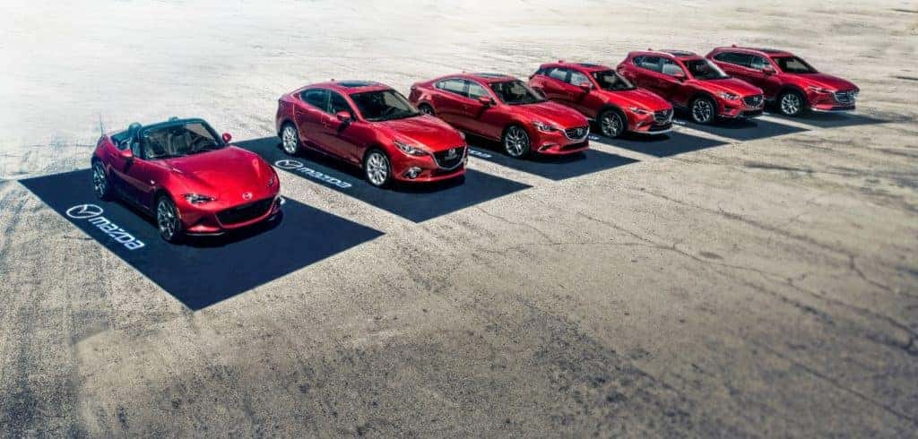 six new red 2020 Mazda cars and SUVs parked on black Mazda-branded car mats on sandy pavement represent the automakers newest lineup for the year
