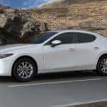 2021 Mazda3 hatchback in a snow-white pearl mica color driving on black asphalt road with a rocky, brown terrain in the background