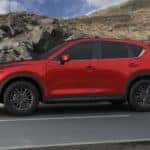Profile shot of a Soul Red Crystal Metallic-colored 2021 Mazda CX-5 Sport trim level on black asphalt road, with a rocky, brown terrain in background