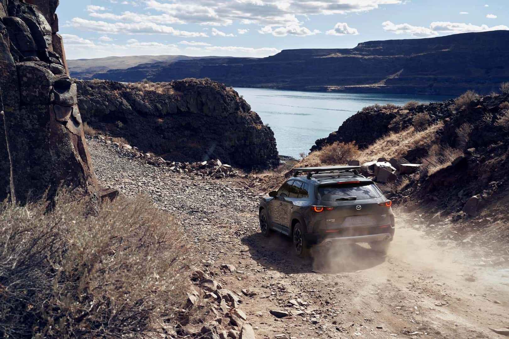 polymetal- gray 2023 Mazda CX-50 driving on a rocky mountain road surrounded by brown cliffs and a blue body of water