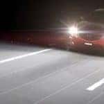 red Mazda driving down a paved road at night with high beams turned on