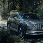 dark gray 2023 Mazda CX-9 driving on a paved road through a forest area