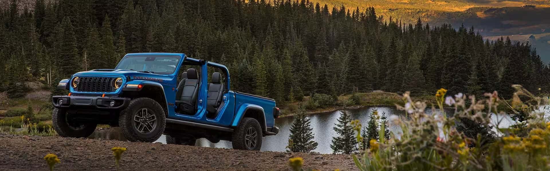 Blue Jeep Gladiator Rubicon with doors removed is parked on a dirt trail overlooking a forested lake and mountain landscape at sunset.