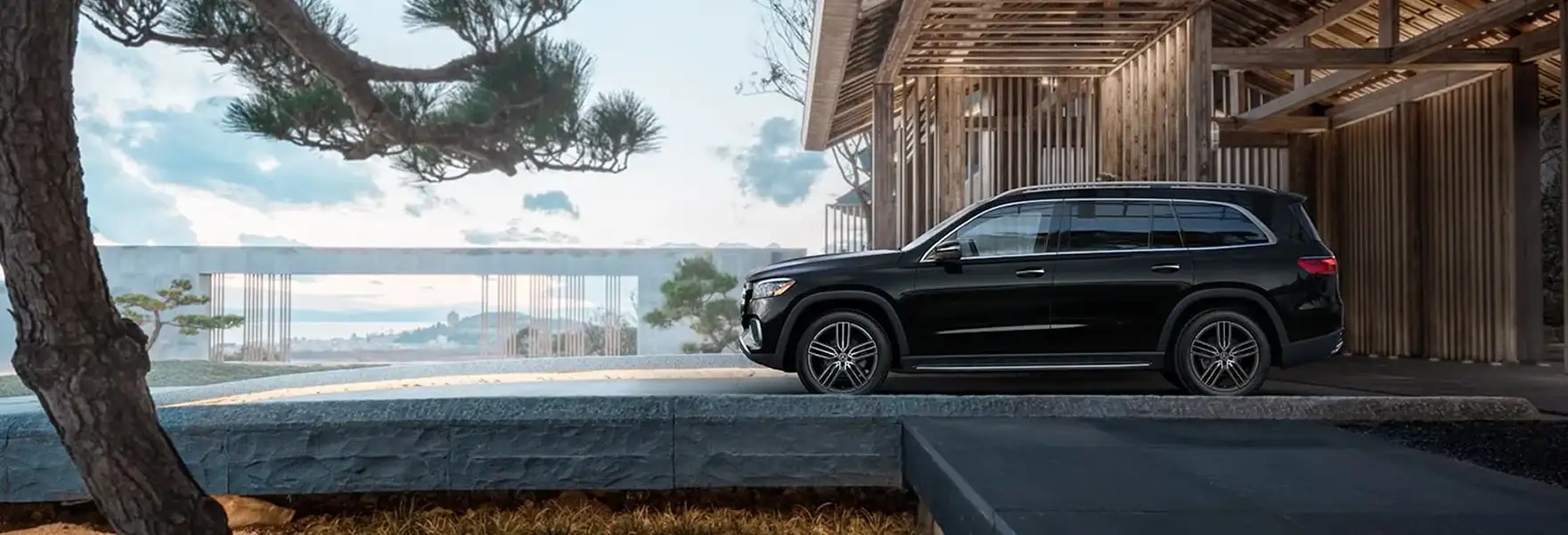 Black Mercedes-Benz GLS SUV parked beside a modern wooden house overlooking the water