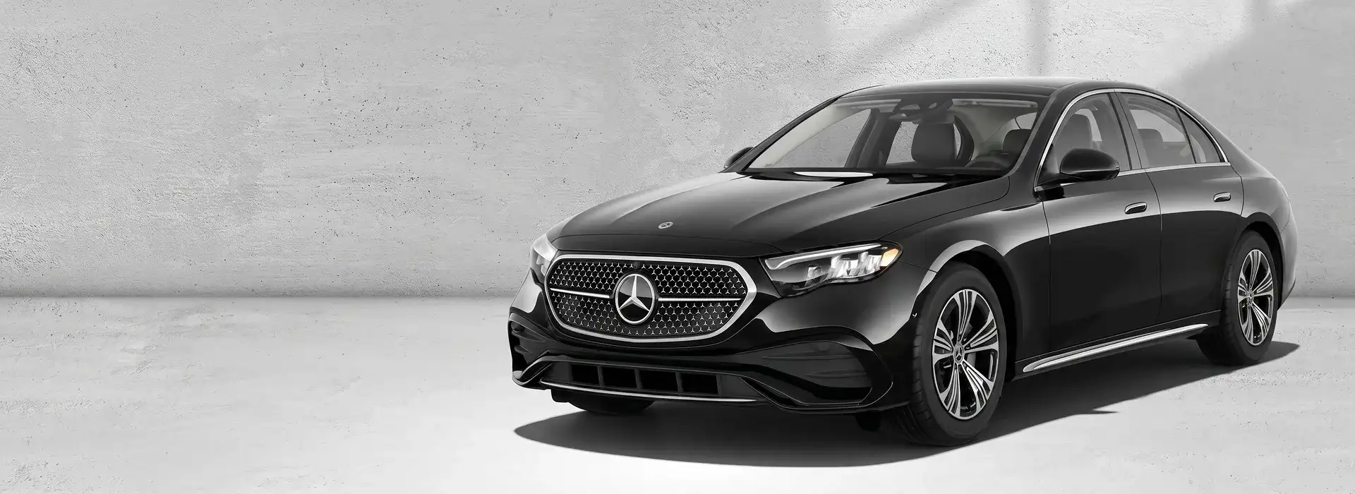 Black 2027 Mercedes-Benz E 350 4MATIC Sedan parked at an angle against a pale concrete wall.