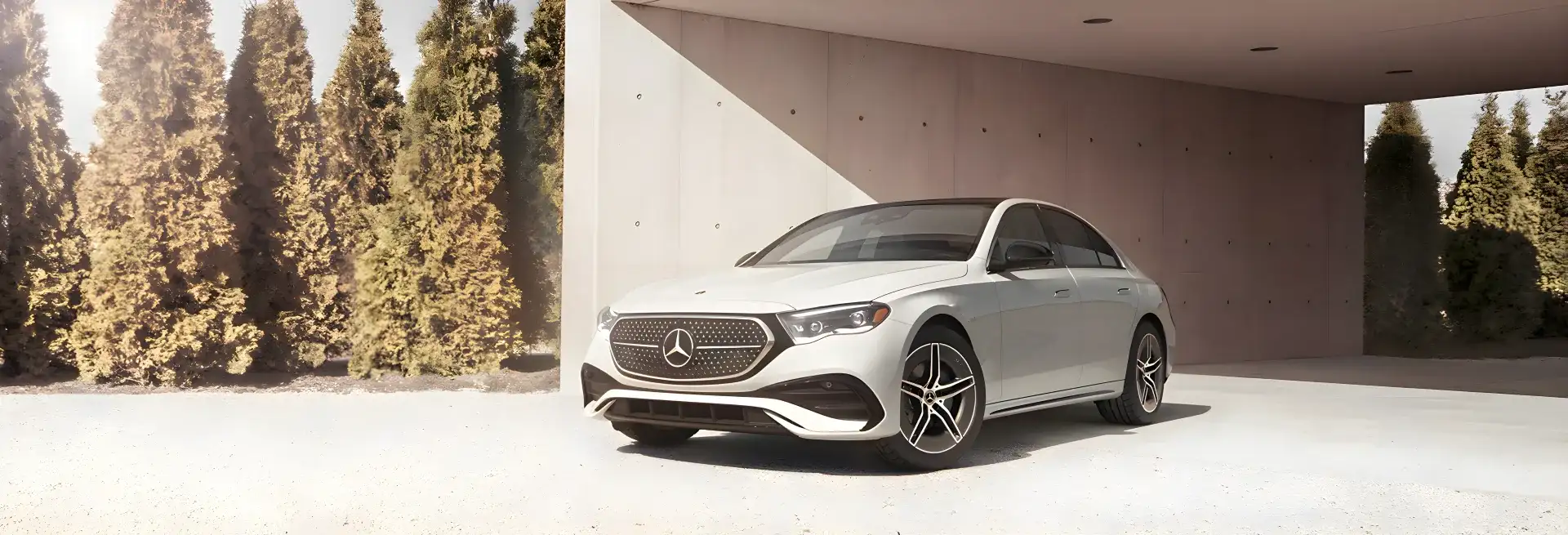 White Mercedes-Benz E 350 4MATIC Sedan parked beneath a concrete carport beside a row of tall evergreens.