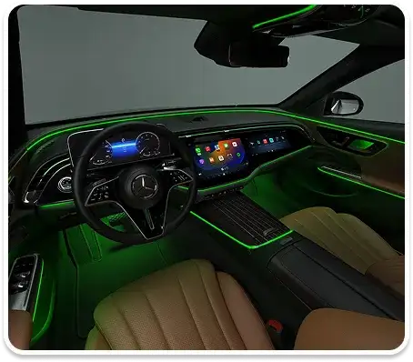 E-Class cabin at night lit in green ambient light, with saddle brown leather seats and dual widescreen displays.