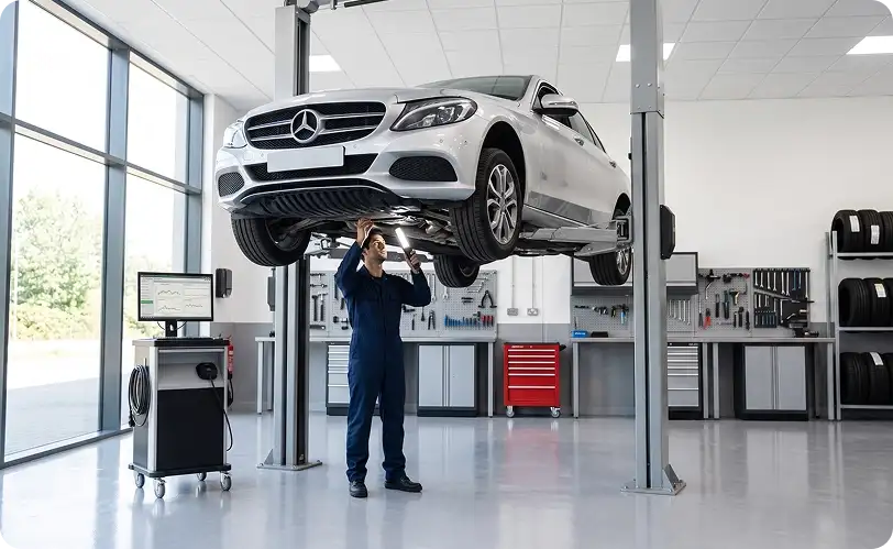 Technician inspecting the underside of a white Mercedes-Benz C-Class raised on a two-post lift