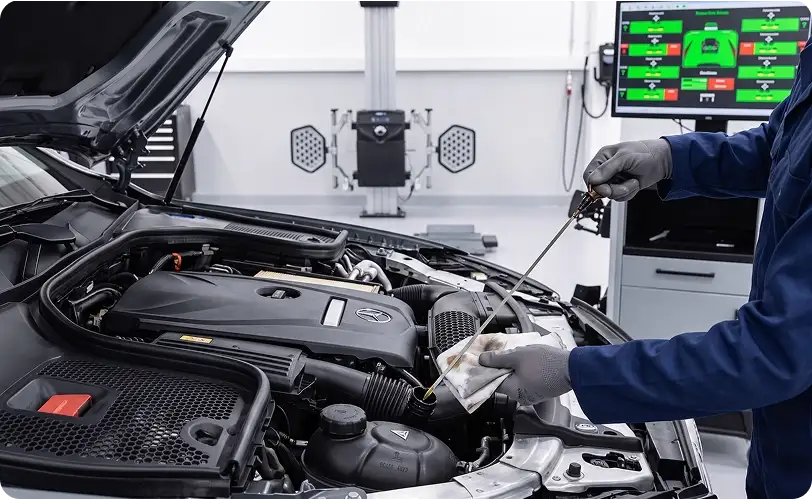 Technician wiping the dipstick of a Mercedes-Benz engine in front of a diagnostic screen showing a vehicle system readout