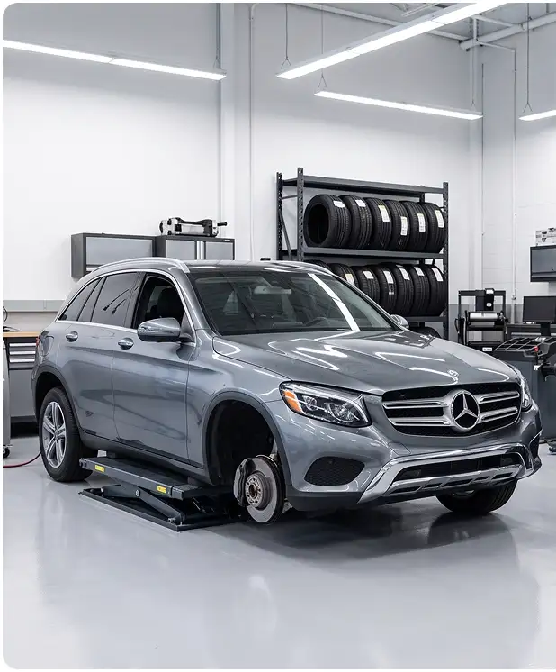 Grey Mercedes-Benz GLC on a low-rise lift with its front wheel removed, in front of a stocked tire rack