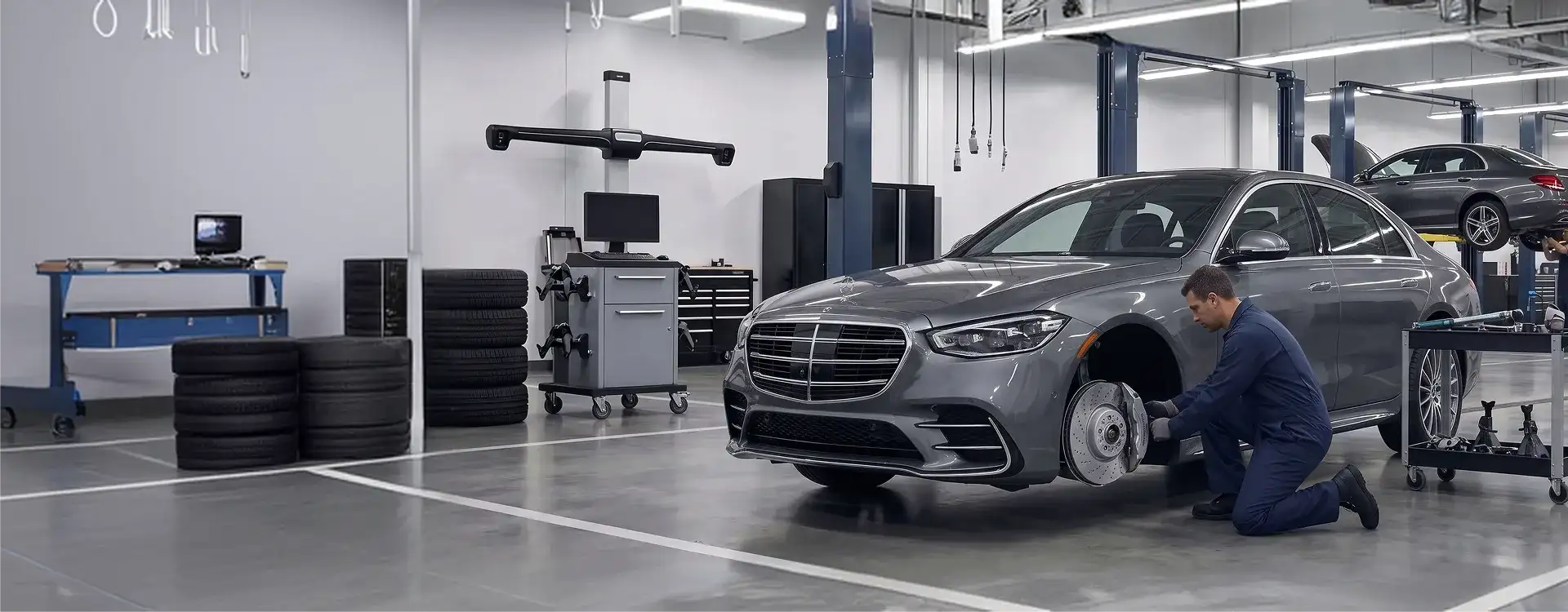 Mercedes-Benz technician kneeling beside a grey Mercedes-Benz S-Class sedan with the front wheel removed inside the Helms Bros service shop