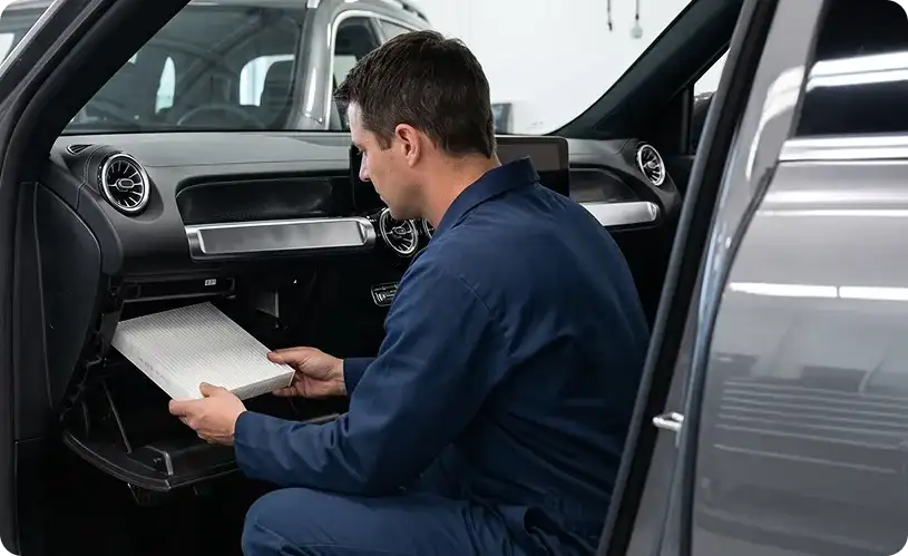 Technician sliding a new cabin filter into place behind the glovebox of a grey Mercedes-Benz GLB