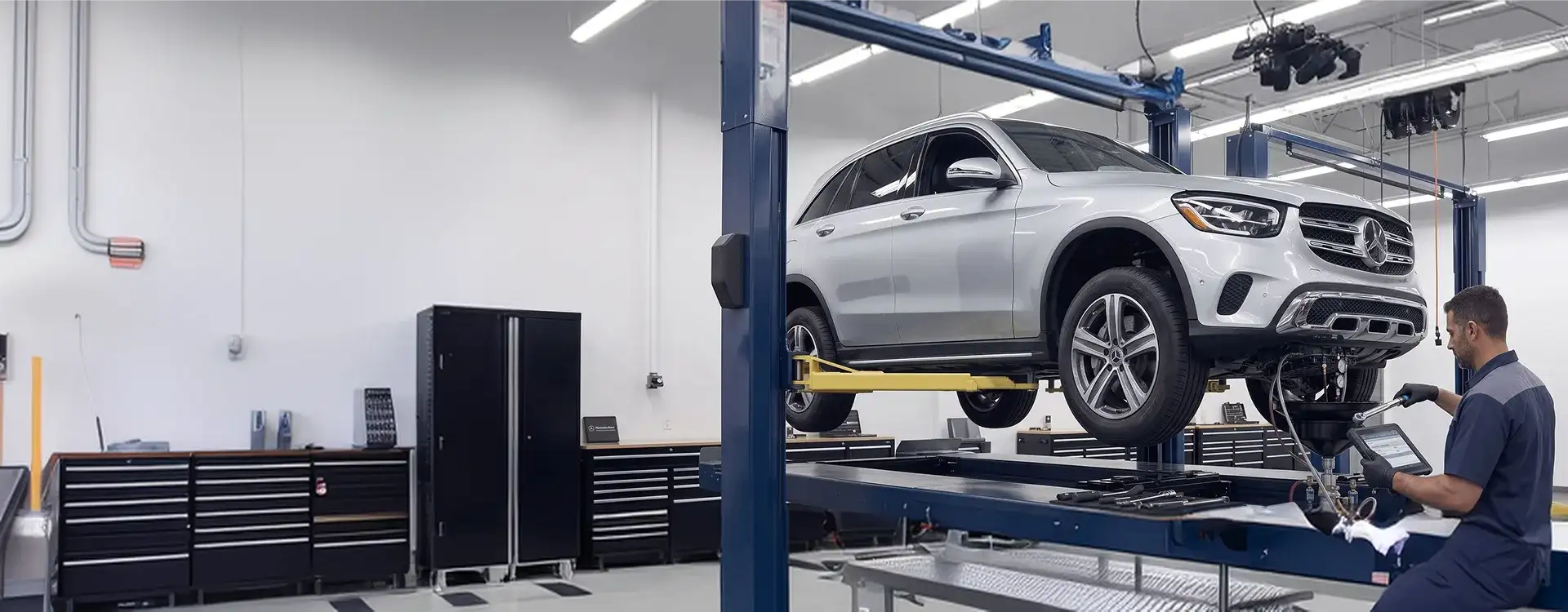 White Mercedes-Benz GLC SUV raised on a two-post lift while a technician reviews transmission data on a diagnostic tablet
