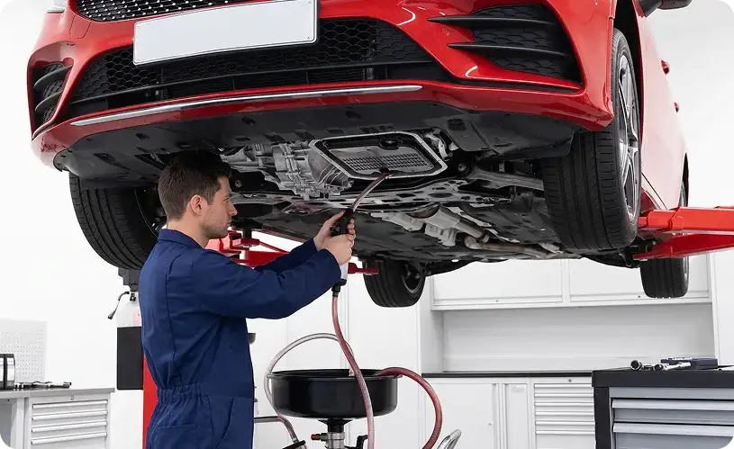 Technician draining transmission fluid from the pan of a red Mercedes-Benz raised on a lift, with a collection drum below