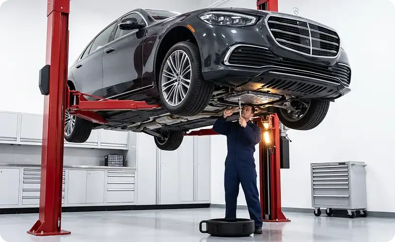 Technician working beneath a dark Mercedes-Benz S-Class raised on a red two-post lift, with a drain pan on the floor