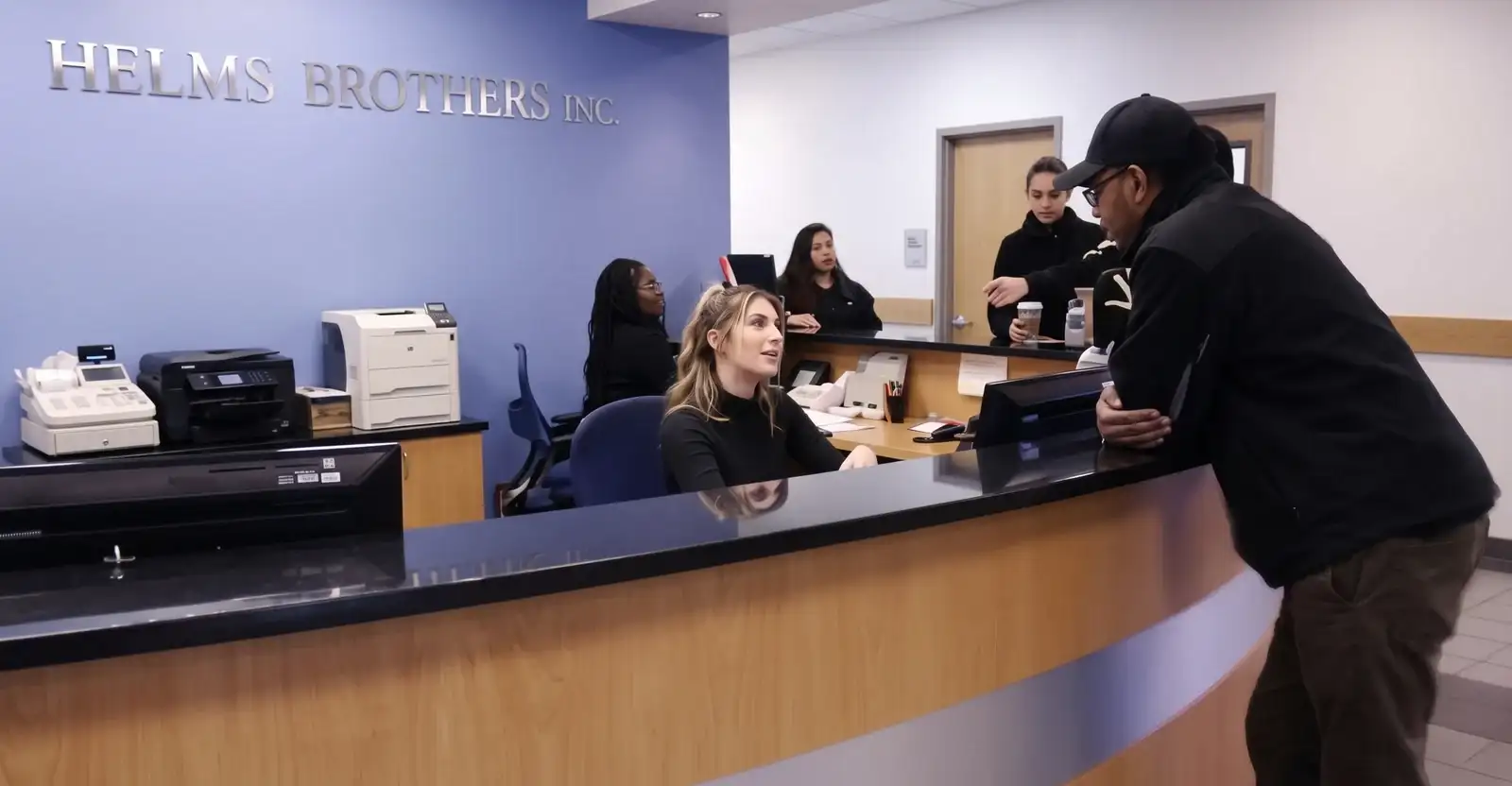 Staff at the Helms Brothers service reception counter serving a customer, the Helms Brothers Inc. sign on the wall behind