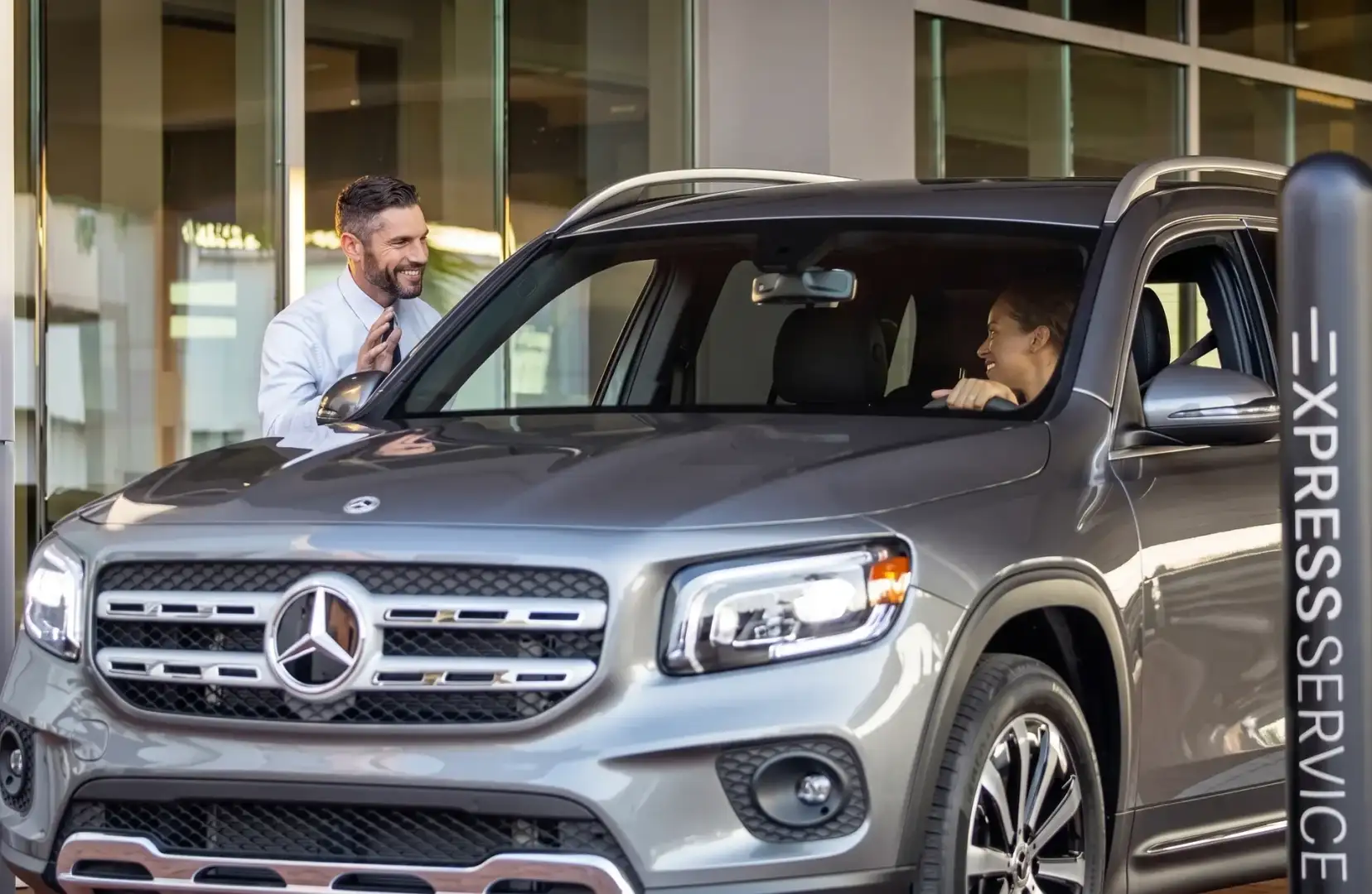 A member of staff in a white shirt leaning in to talk to a driver through the window of a silver Mercedes-Benz SUV outside the dealership