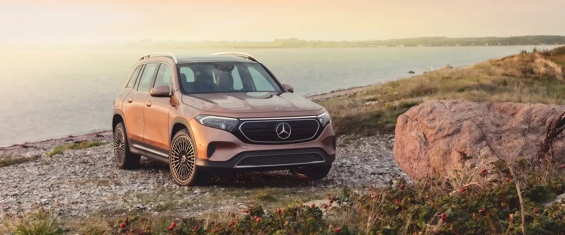 A copper Mercedes-Benz electric SUV parked on a rocky shoreline at sunset, front three-quarter view