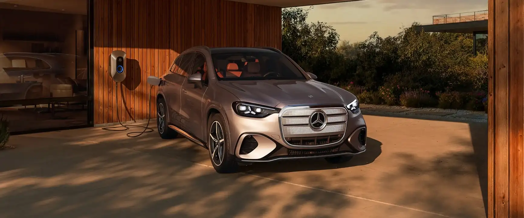 A silver Mercedes-Benz GLC electric SUV plugged into a wall charger under a timber carport at sunset