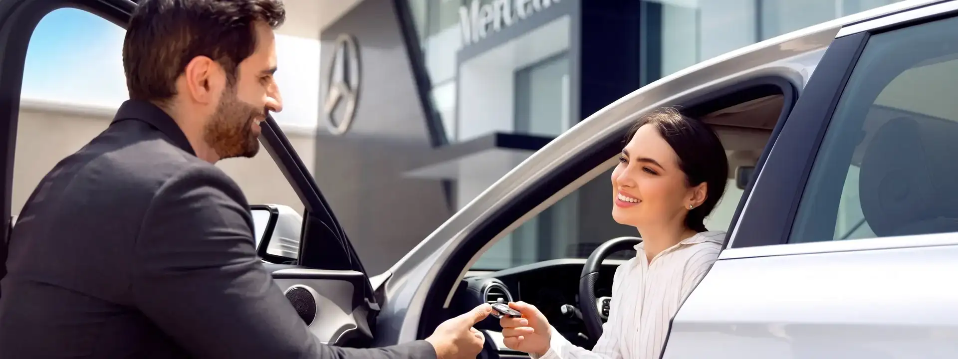 A salesperson in a dark suit hands a key fob to a smiling woman in the driver seat of a white Mercedes-Benz, the dealership star sign on the building behind