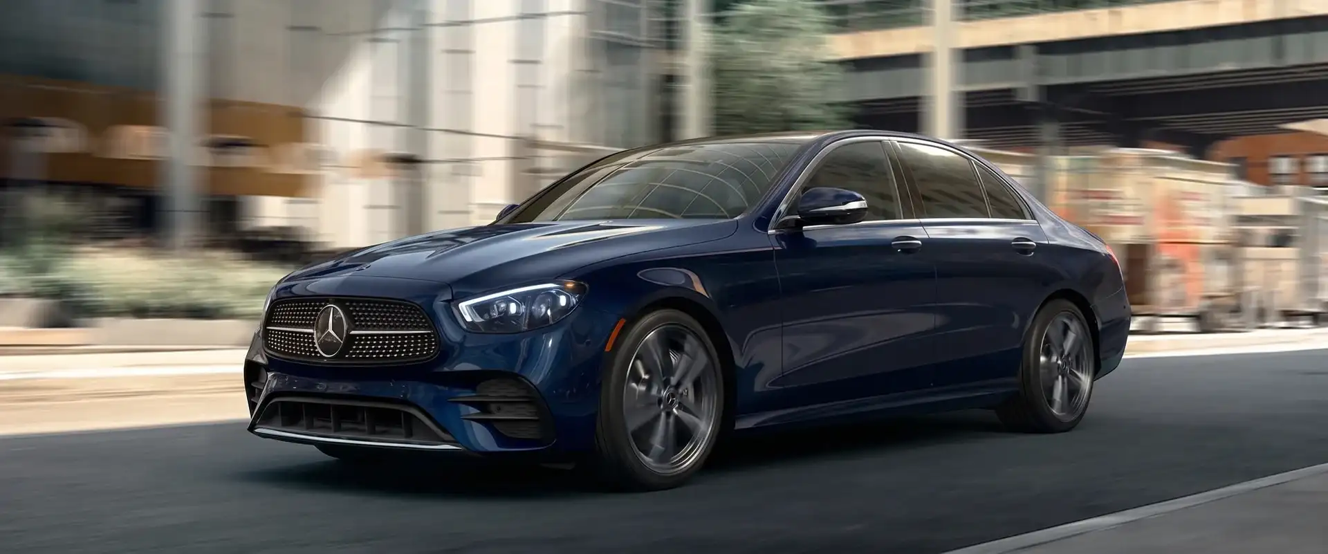 A dark blue Mercedes-Benz sedan driving a city street past glass buildings, front three-quarter view