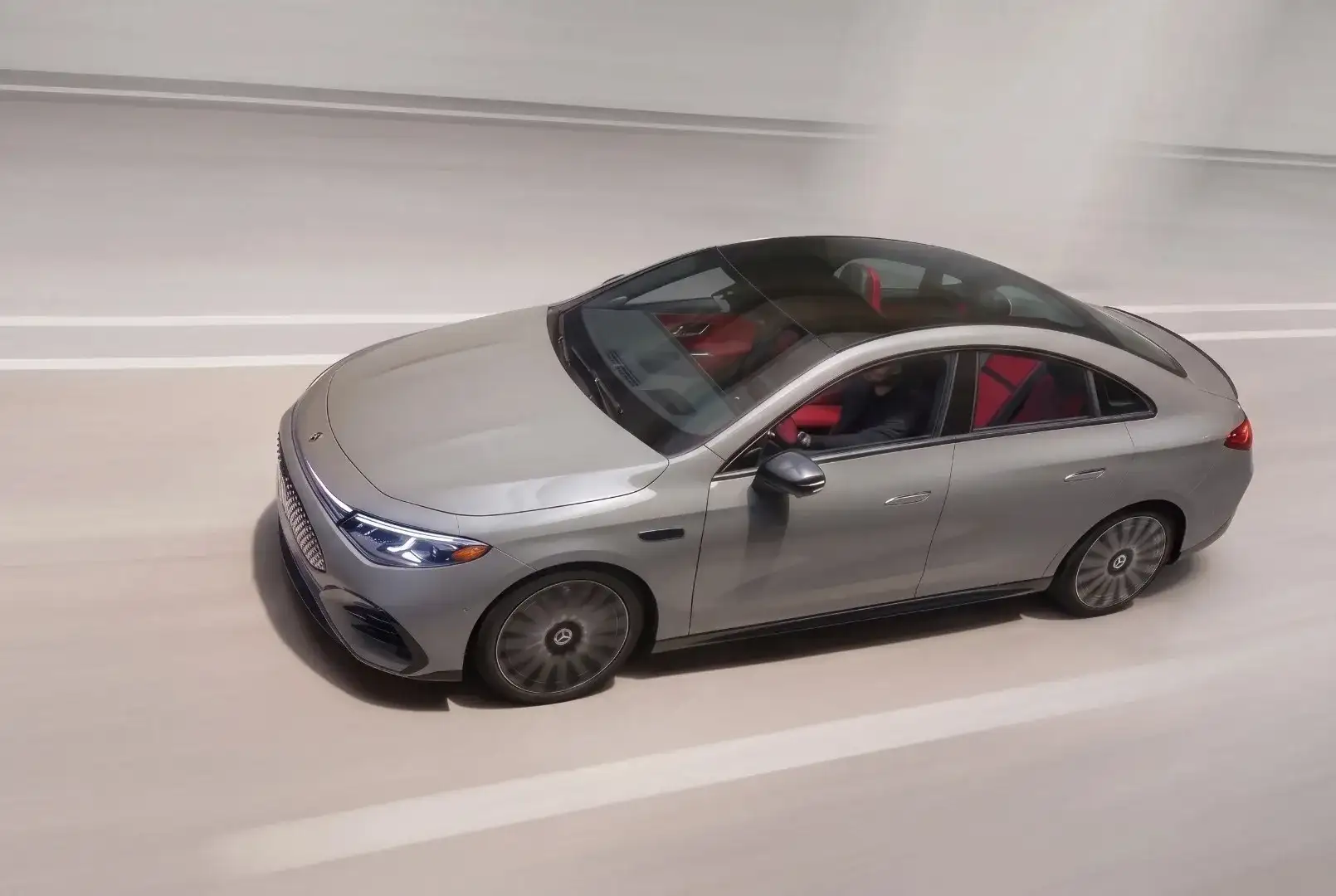 A silver Mercedes-Benz sedan with a panoramic roof and red interior, driving a pale road seen from above.