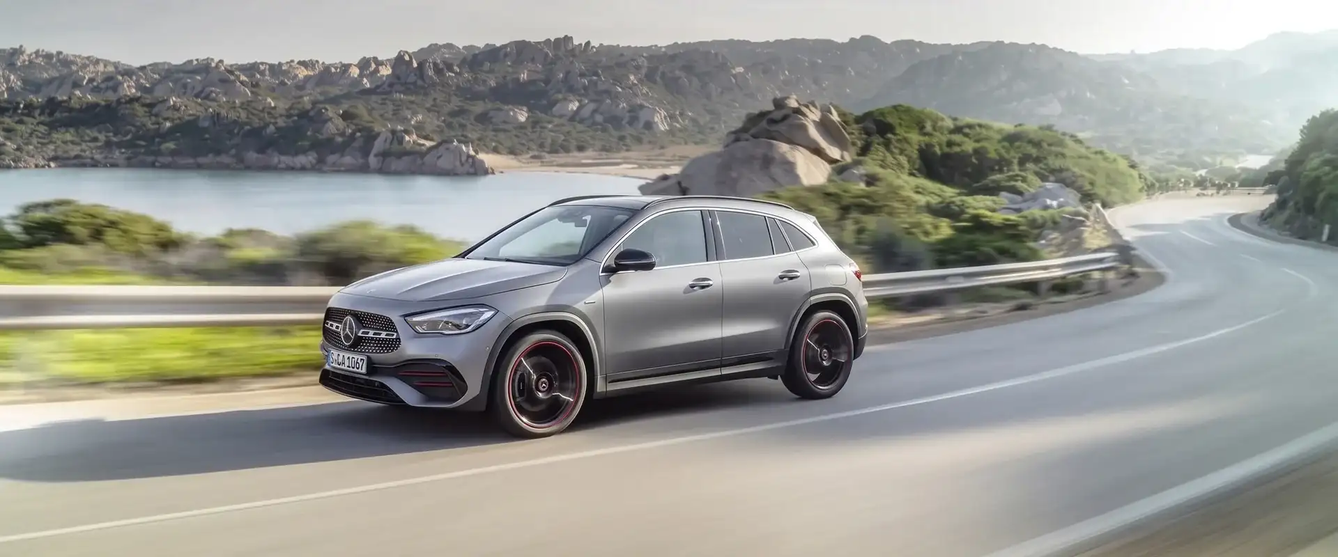 A grey Mercedes-Benz SUV driving a coastal road past a rocky bay, front three-quarter view