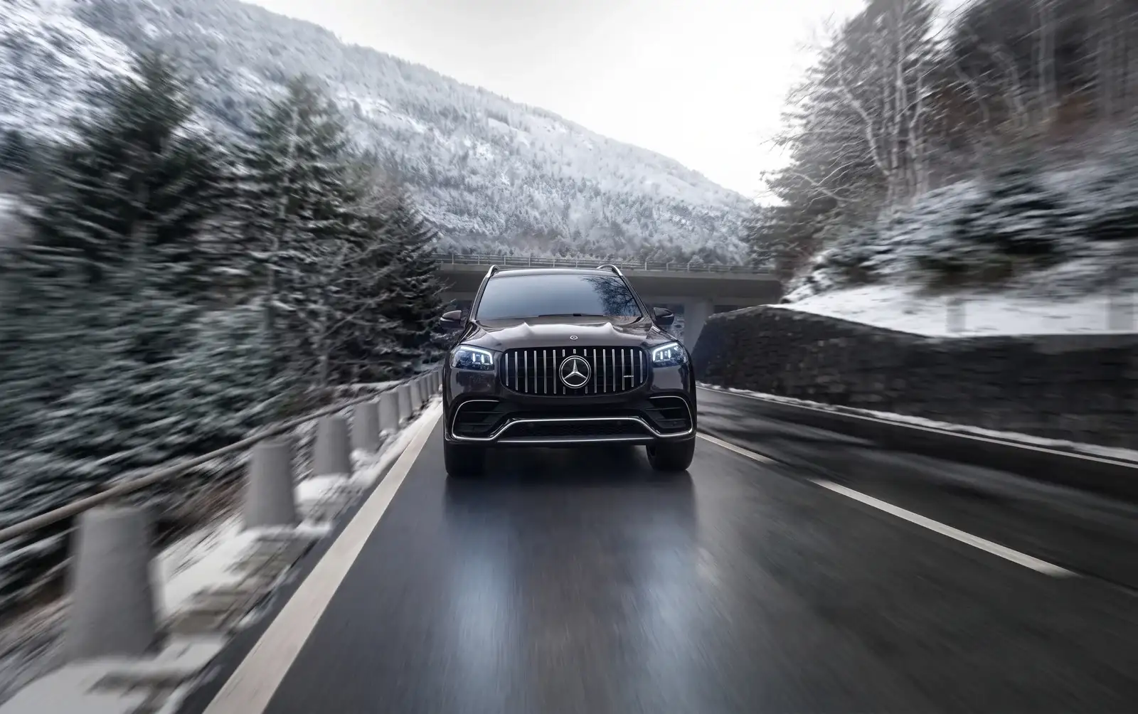 A dark Mercedes-Benz SUV driving head-on along a snowy mountain road, pine trees and a bridge behind