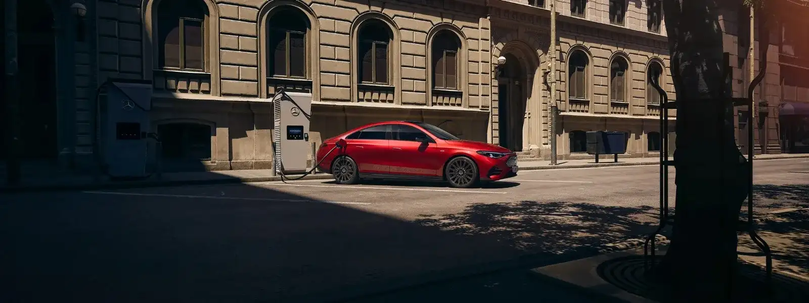A red Mercedes-Benz electric sedan plugged into a Mercedes-Benz public charging station on a city street