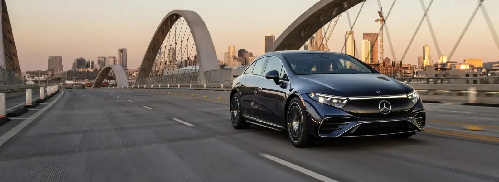 A dark blue Mercedes-Benz electric sedan crossing a city bridge at dusk, the downtown skyline behind