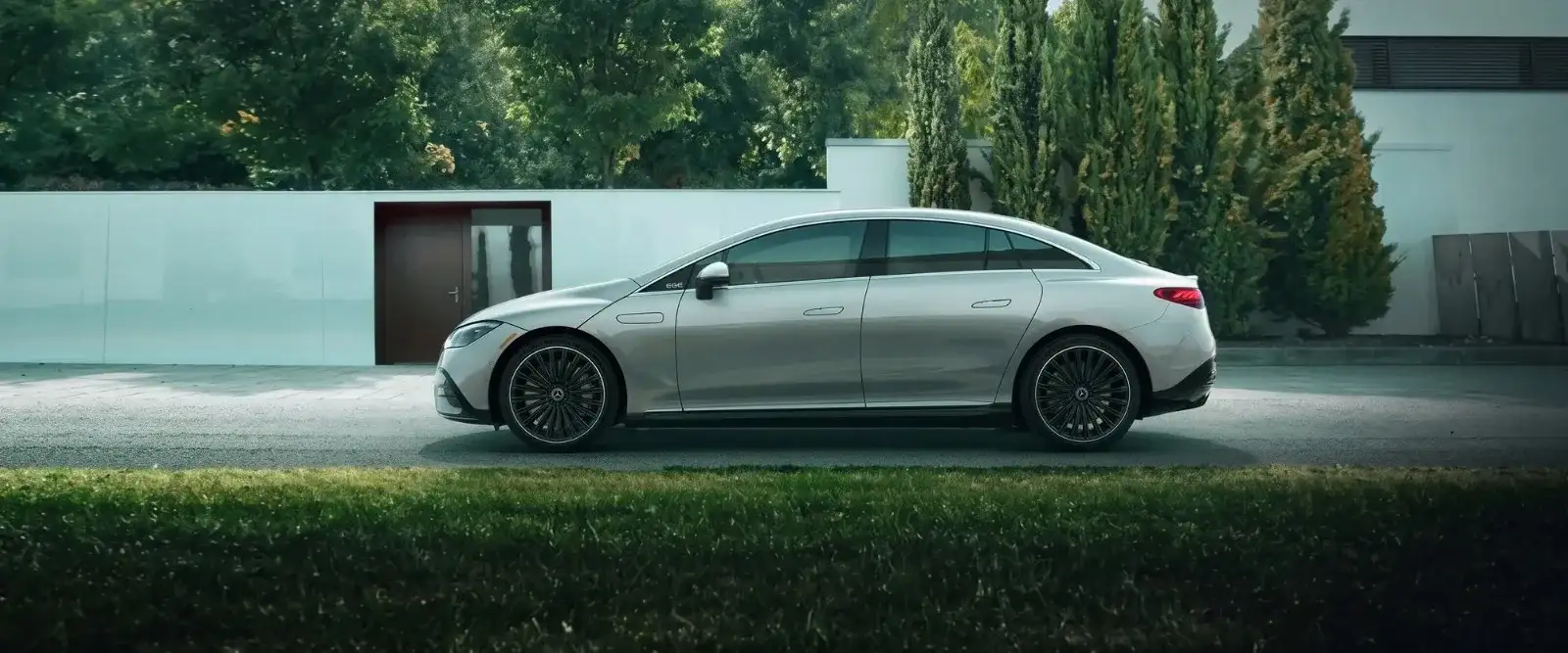 A silver Mercedes-Benz EQE sedan in side profile on the drive of a modern house, cypress trees behind
