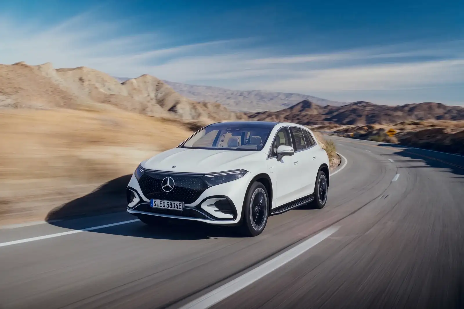 A white Mercedes-Benz EQS SUV driving a desert mountain road, front three-quarter view