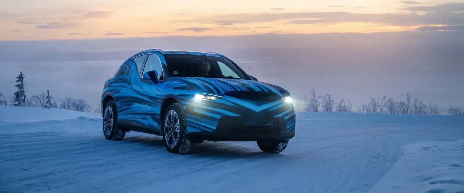 An SUV in blue and black test camouflage on a snow-covered road at dusk, headlights lit