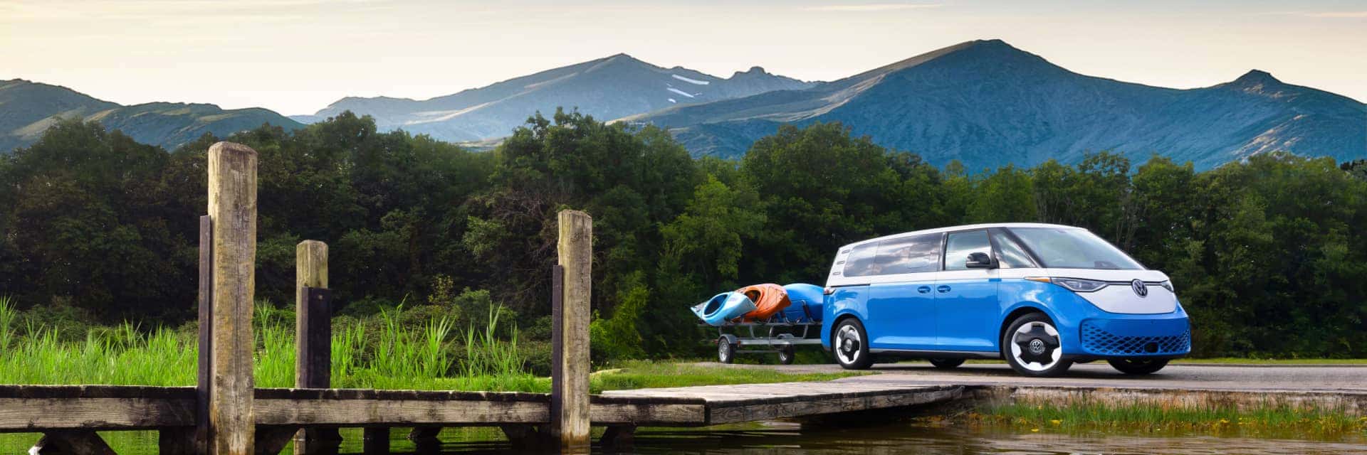 ID.Buzz parked along a dock on a lake with kayaks hitched to the back with mountain background.