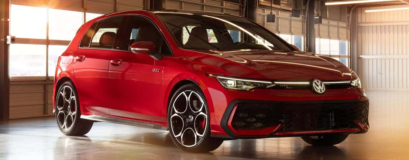 Red 2026 Volkswagen Golf GTI hatchback parked in a sunlit garage, featuring a glowing front VW emblem and black machined alloy wheels.