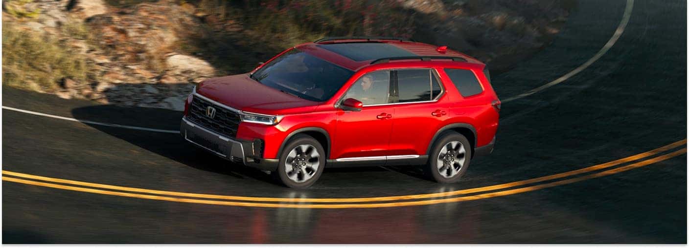 A red 2026 Honda Pilot SUV navigates a sharp curve on an asphalt road, set against a backdrop of a rocky hillside.
