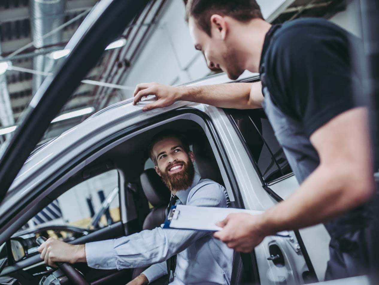 Man in a suit sits inside a car and smiling at a mechanic who holds a clipboard.