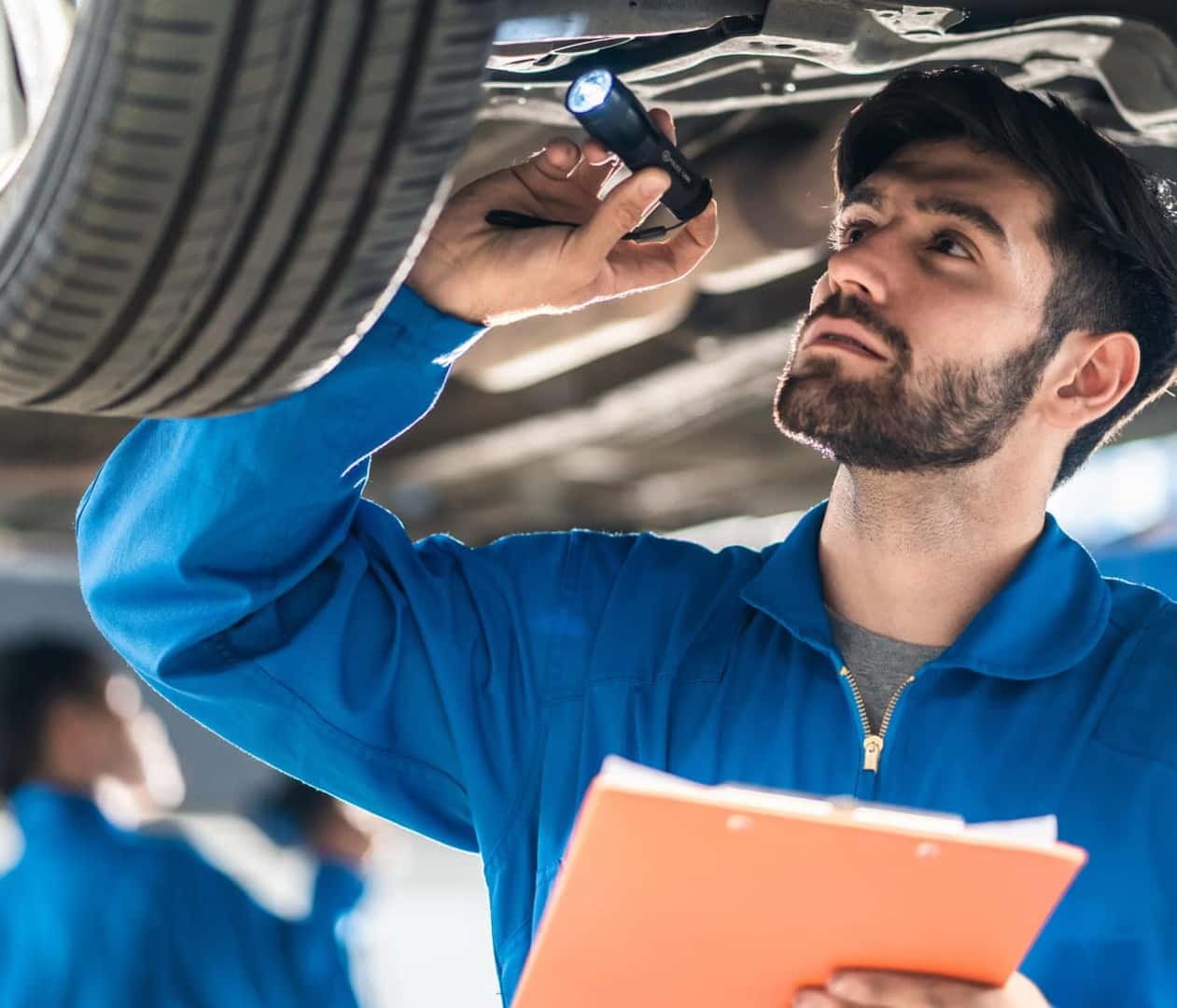 Mechanic in a blue uniform inspects a car's underside using a flashlight and holding a clipboard.