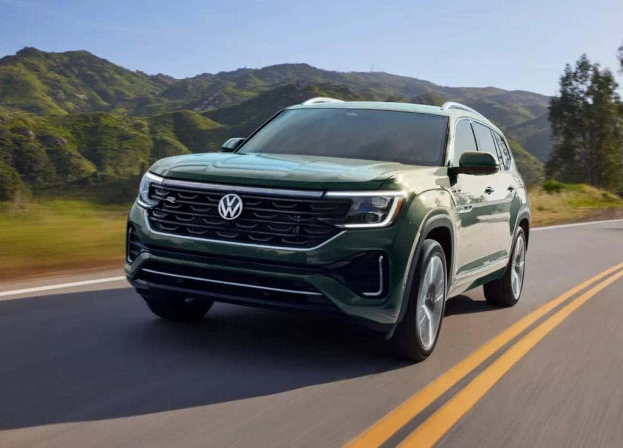 2026 Volkswagen Atlas driving on a winding road flanked by lush green hills under a clear blue sky.