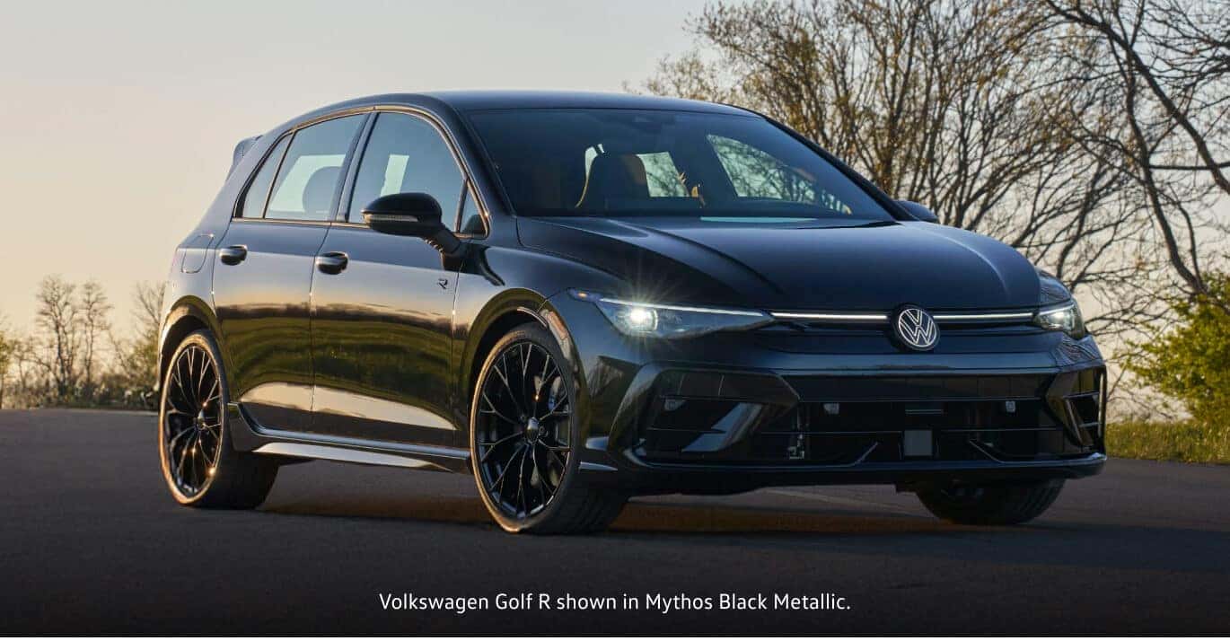 2026 Volkswagen Mythos Black Metallic Volkswagen Golf R parked on a paved road with trees in the background.