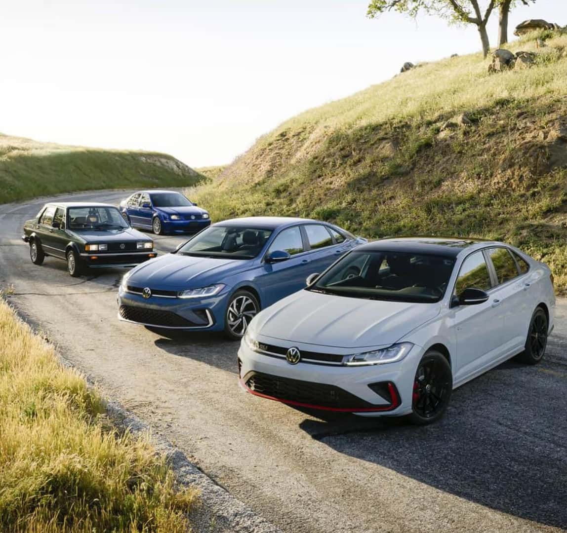Several VW Jetta cars from each model generation parked side-by-side on a sunny hillside road.