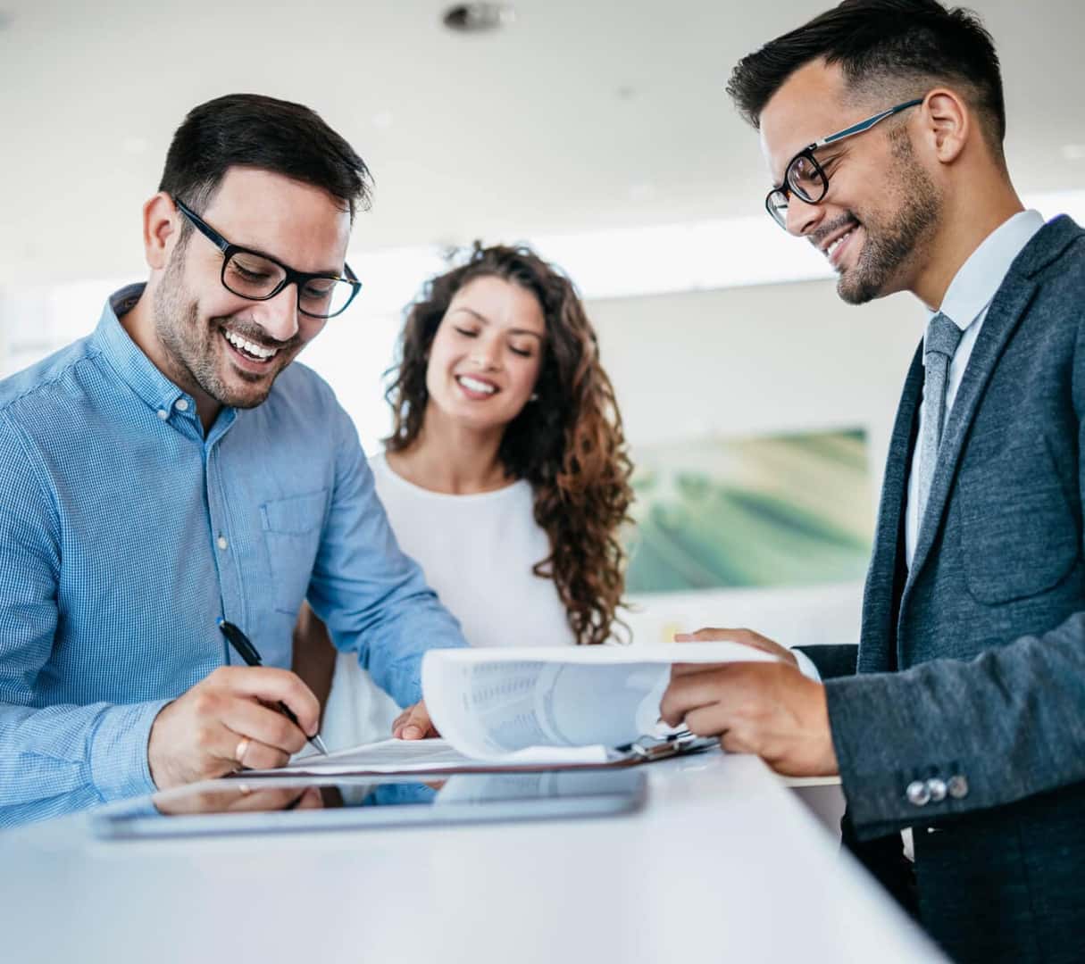 A sales associate assists a smiling couple handling financing paperwork inside a sunny car dealership.